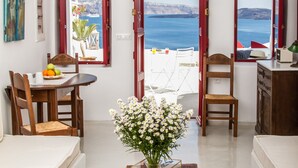A room with a view of the sea, a dining area with a table and chairs, a wooden cabinet, and a vase with flowers. - Hector Cave House (Santorini)