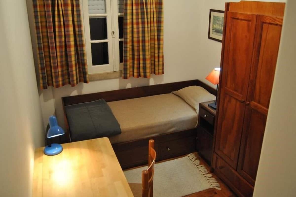 Quarto pequeno e funcional, com uma cama de solteiro, escrivaninha, cadeira, guarda-roupa e janela com cortinas.
