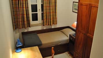 Une petite pièce, de taille modeste, comprenant un lit simple, un bureau, une chaise, une armoire et une fenêtre avec des rideaux.