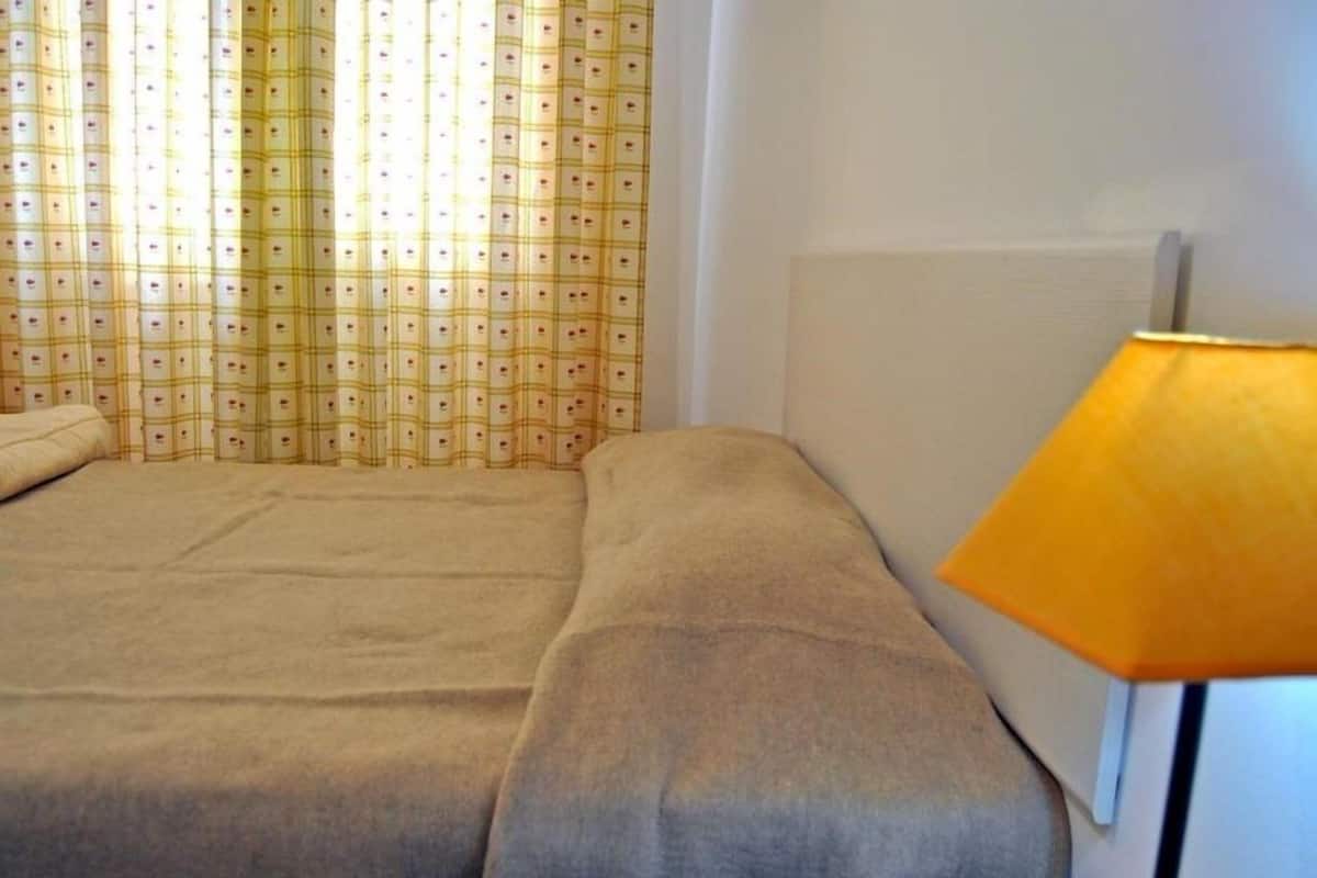 Uma cama de solteiro com um cobertor bege, uma mesinha de cabeceira de madeira e um abajur amarelo.
