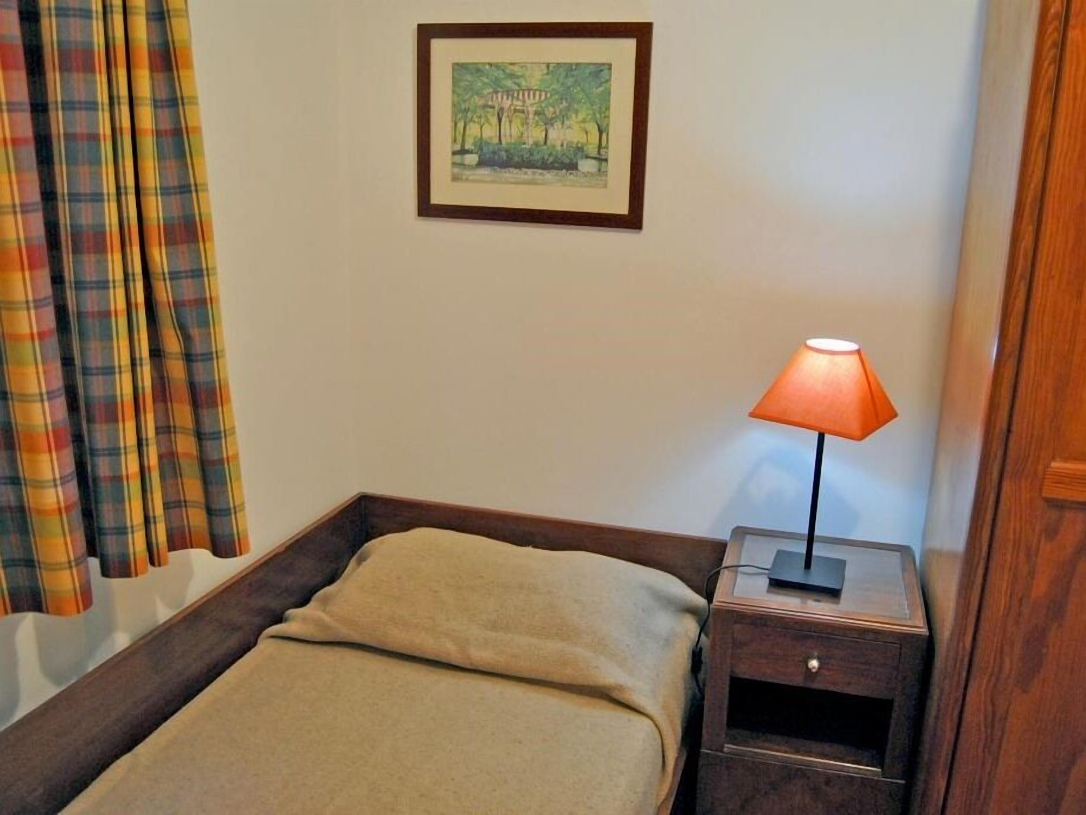 A single bed with a beige blanket, a nightstand with a lamp, a framed picture on the wall, and plaid curtains.