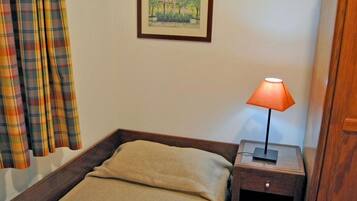 A single bed with a beige blanket, a nightstand with a lamp, a framed picture on the wall, and plaid curtains.