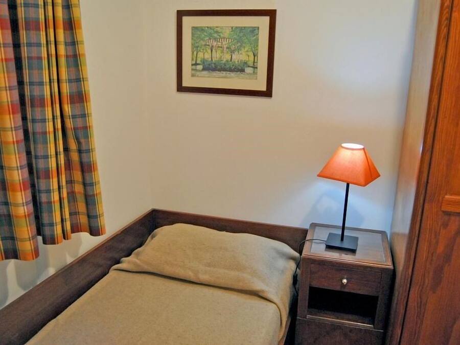 A single bed with a beige blanket, a nightstand with a lamp, a framed picture on the wall, and plaid curtains.