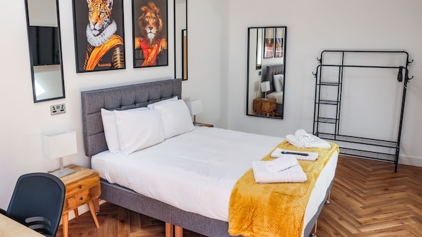 Habitación de hotel ordenada con cama, escritorio y silla. En la pared hay cuadros enmarcados, un espejo y una estructura metálica.