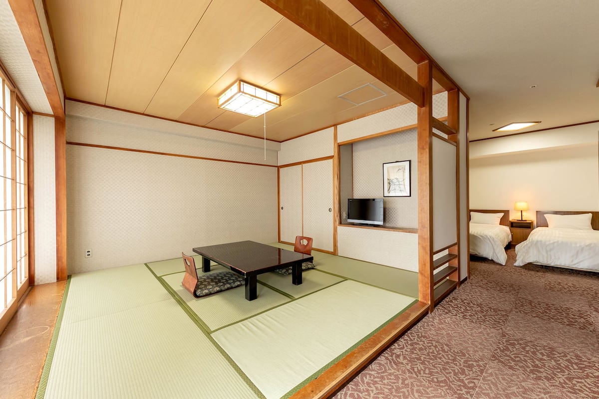 Ett traditionellt japanskt rum med tatamimattor på golvet, ett lågt bord, två sängar, en TV och ett nattduksbord med en lampa.