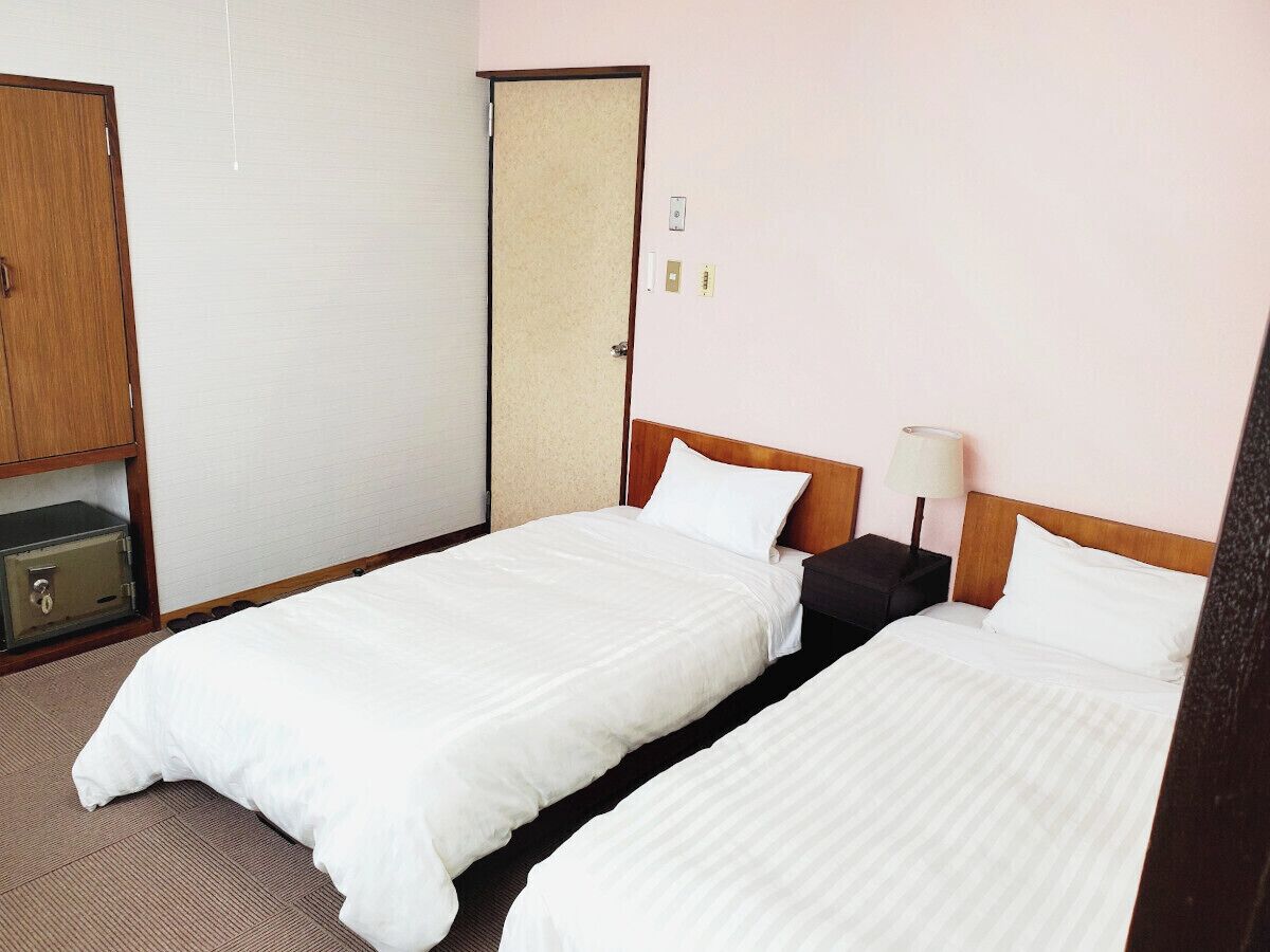 A hotel room with two single beds, a wooden headboard, a bedside table with a lamp, and a safe.