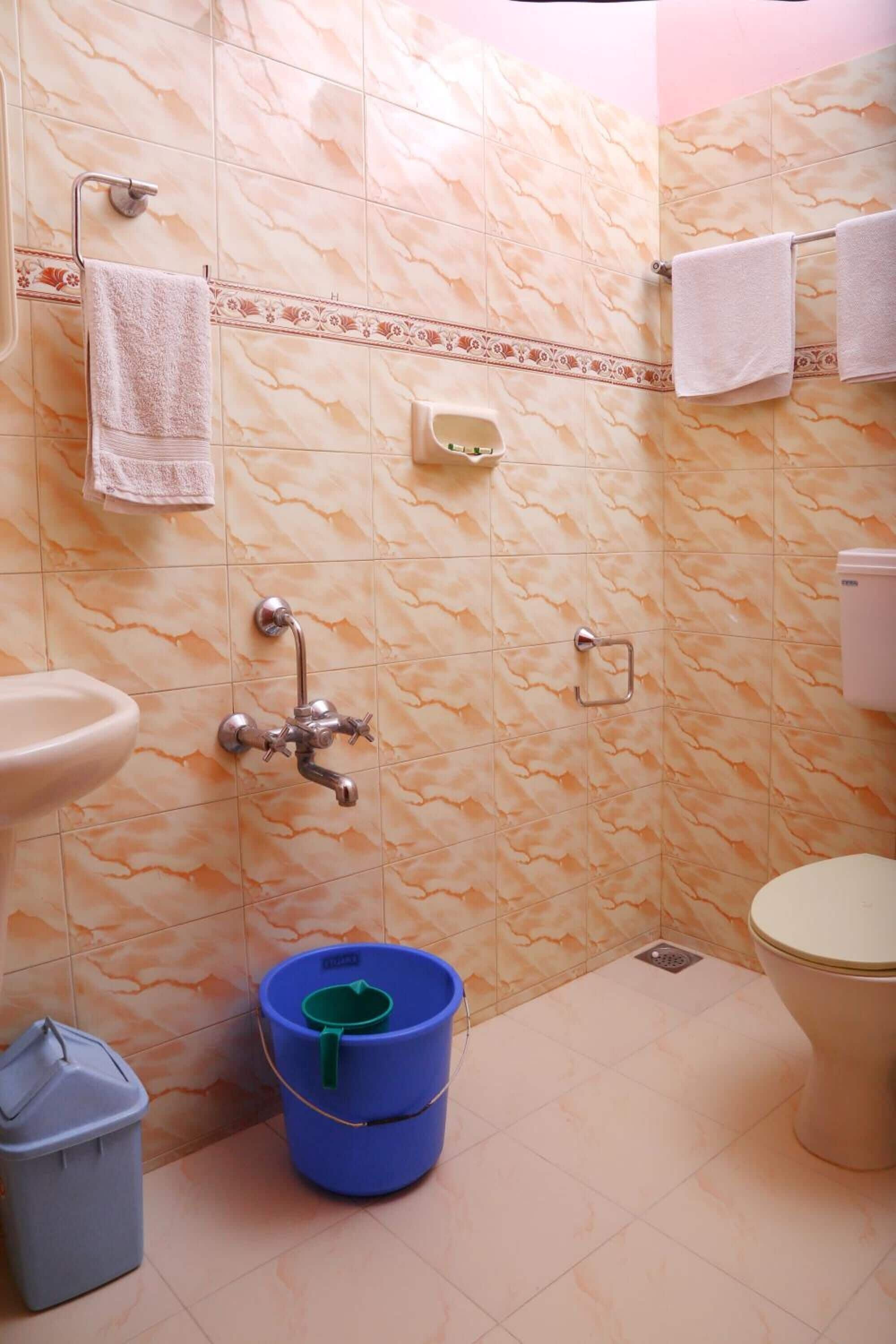 Une salle de bain carrelée de marbre, avec un lavabo, des toilettes, une douche et un seau bleu.