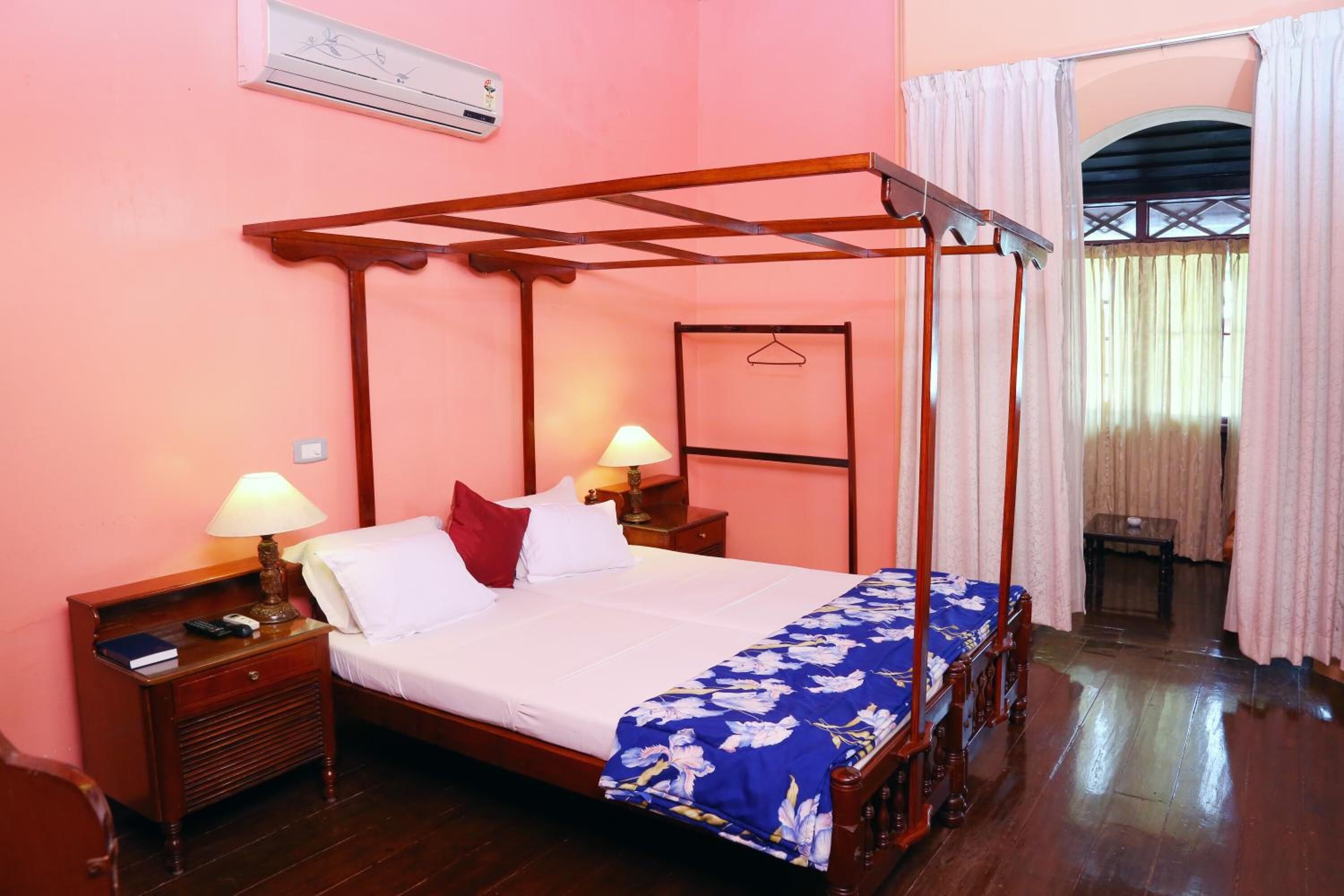 A four-poster bed with a wooden frame, white bedding, and a blue floral throw. There are two bedside tables with lamps, a hanging coat rack, and a window with curtains.