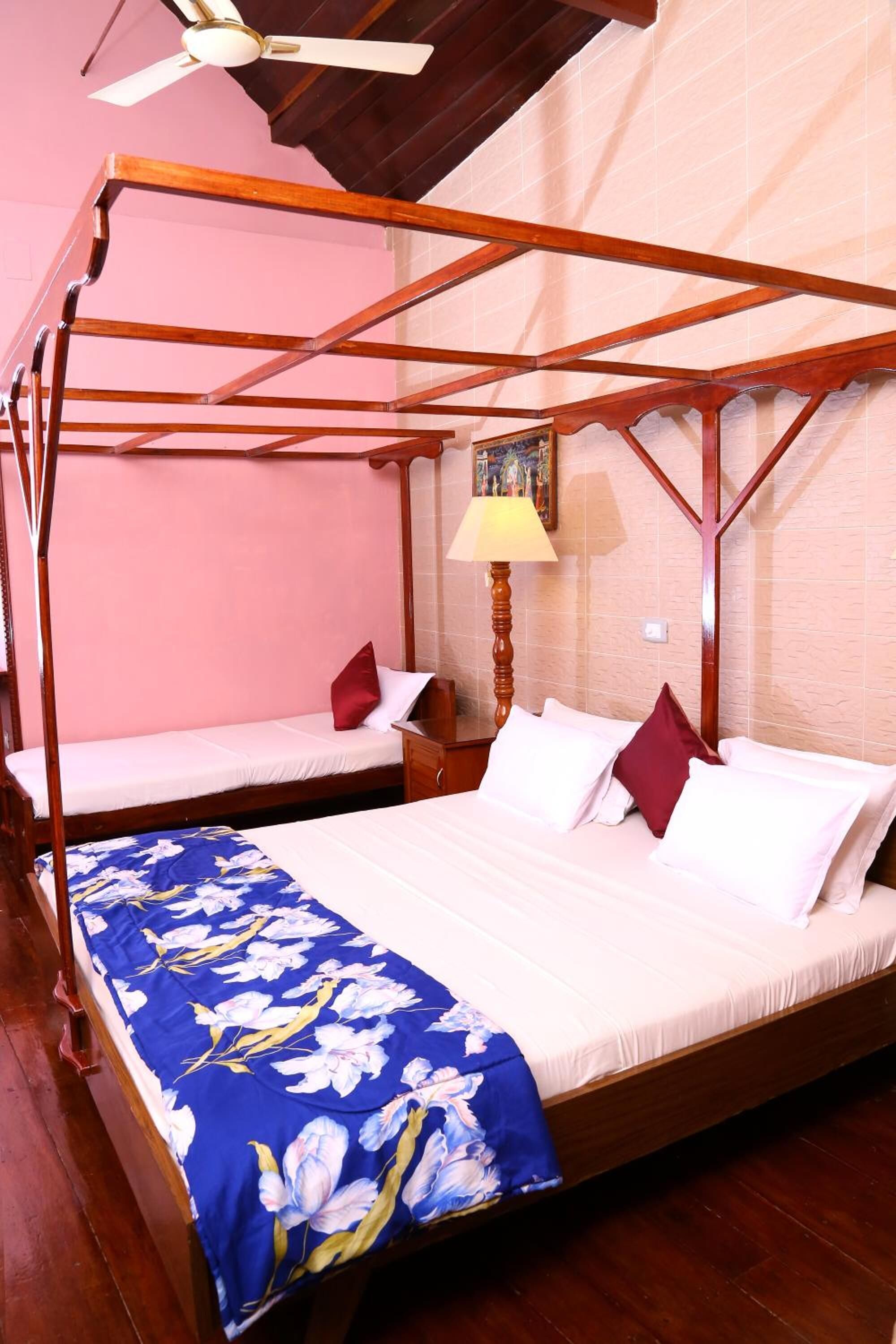 A room with a four-poster bed, a smaller bed, wooden flooring, a ceiling fan, and a lamp.