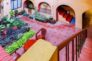 Um pátio com piso de telhas vermelhas, plantas em vasos, uma mesa de madeira e uma escadaria com degraus vermelhos.
