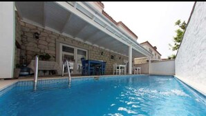 Una piscina interior con agua cristalina azul, una pared de piedra y un área de patio techada con una mesa y sillas azules.