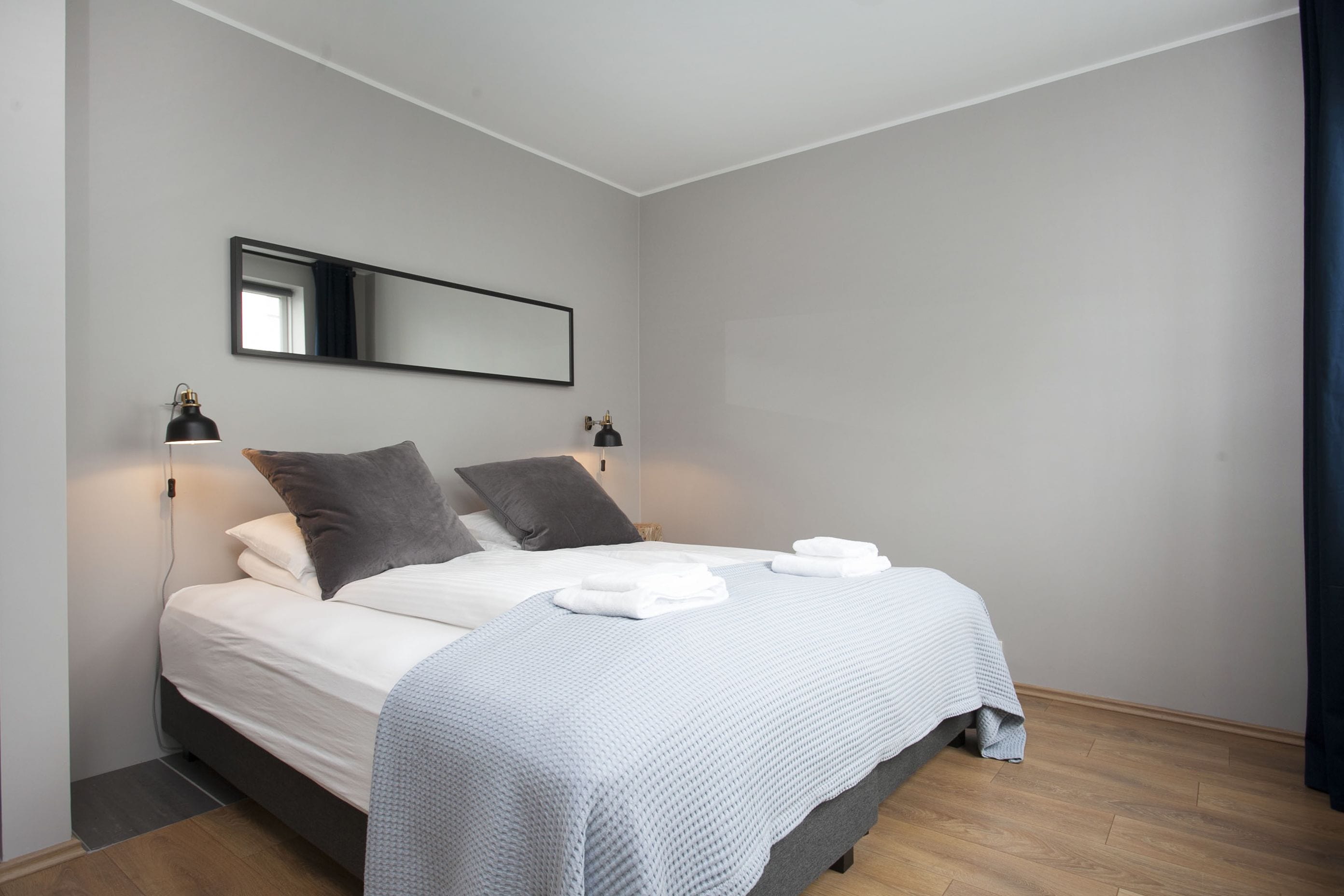 Een net opgemaakt bed met witte lakens en een grijze deken, twee bedlampen en een grote spiegel aan de muur.