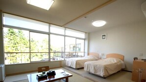 Une chambre d’hôtel avec un sol recouvert de tatamis, deux lits, un bureau avec une chaise et une grande fenêtre donnant sur l’extérieur.