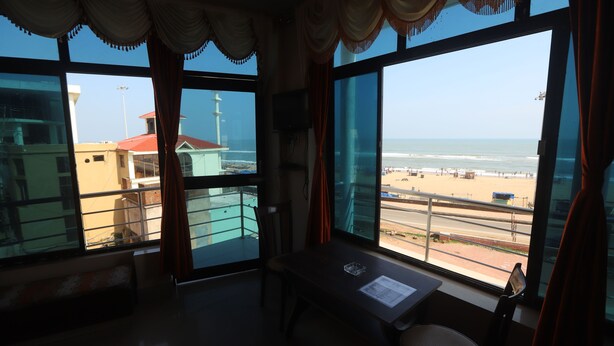 A room with a large window overlooking a beach, a table with a chair, and a view of buildings and a lighthouse. - Larica Holiday Inn Puri (Puri)