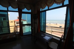 A room with a large window overlooking a beach, a table with a chair, and a view of buildings and a lighthouse.