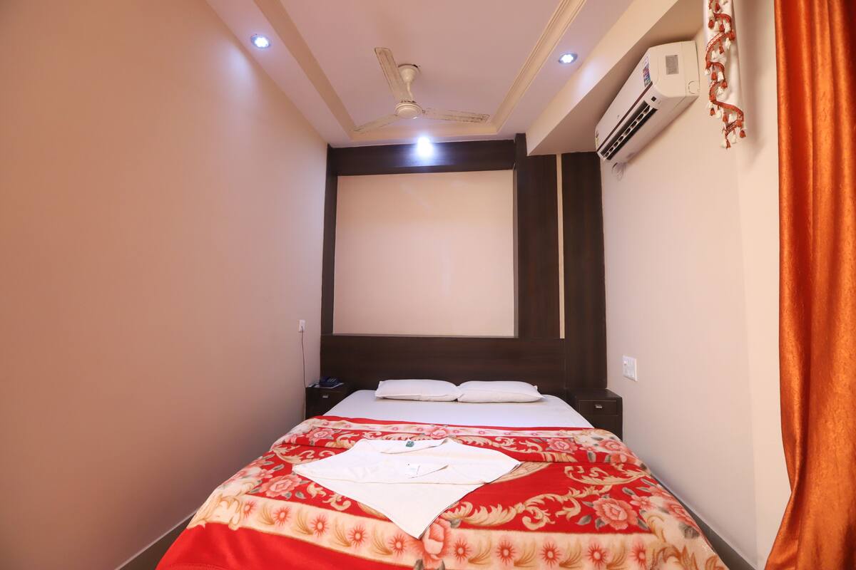 A hotel room with a bed, a ceiling fan, an air conditioning unit, and a window with curtains.