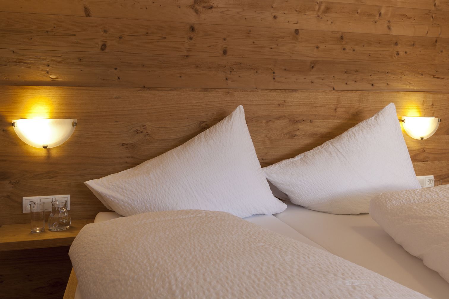 A neatly made bed with white bedding and pillows, wooden headboard, and two wall-mounted light fixtures.