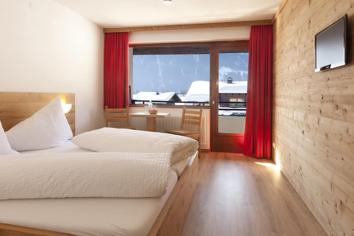 A hotel room with a bed, a small table, a chair, and a view of a snowy landscape through a glass door.