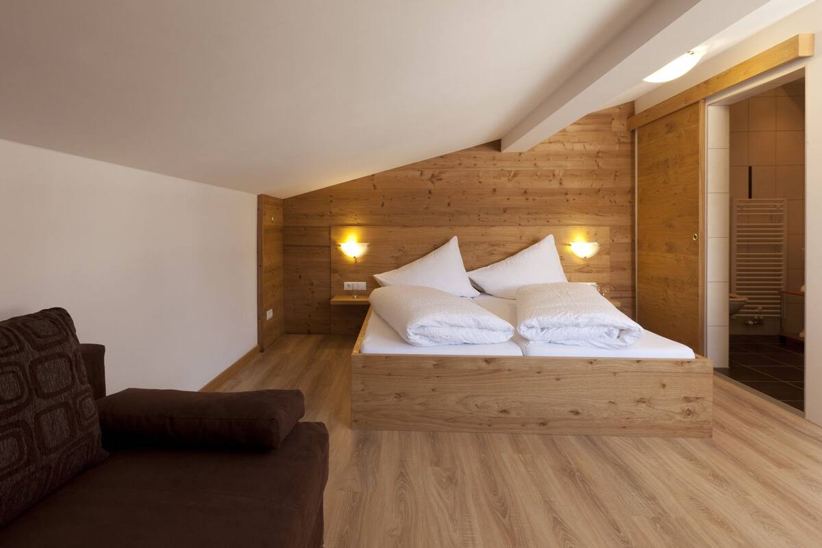 A modern bedroom with a wooden ceiling and walls, a bed with white linens, a nightstand with a lamp, and a brown sofa.