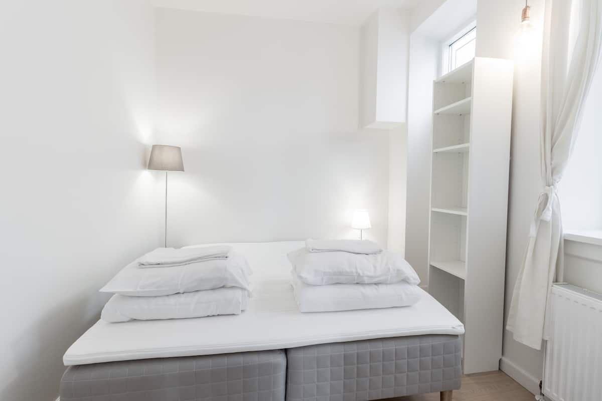 Ein ordentlich gemachtes Bett mit Kissen, einer Nachttischlampe und einem weißen Kleiderschrank mit geöffneten Türen.