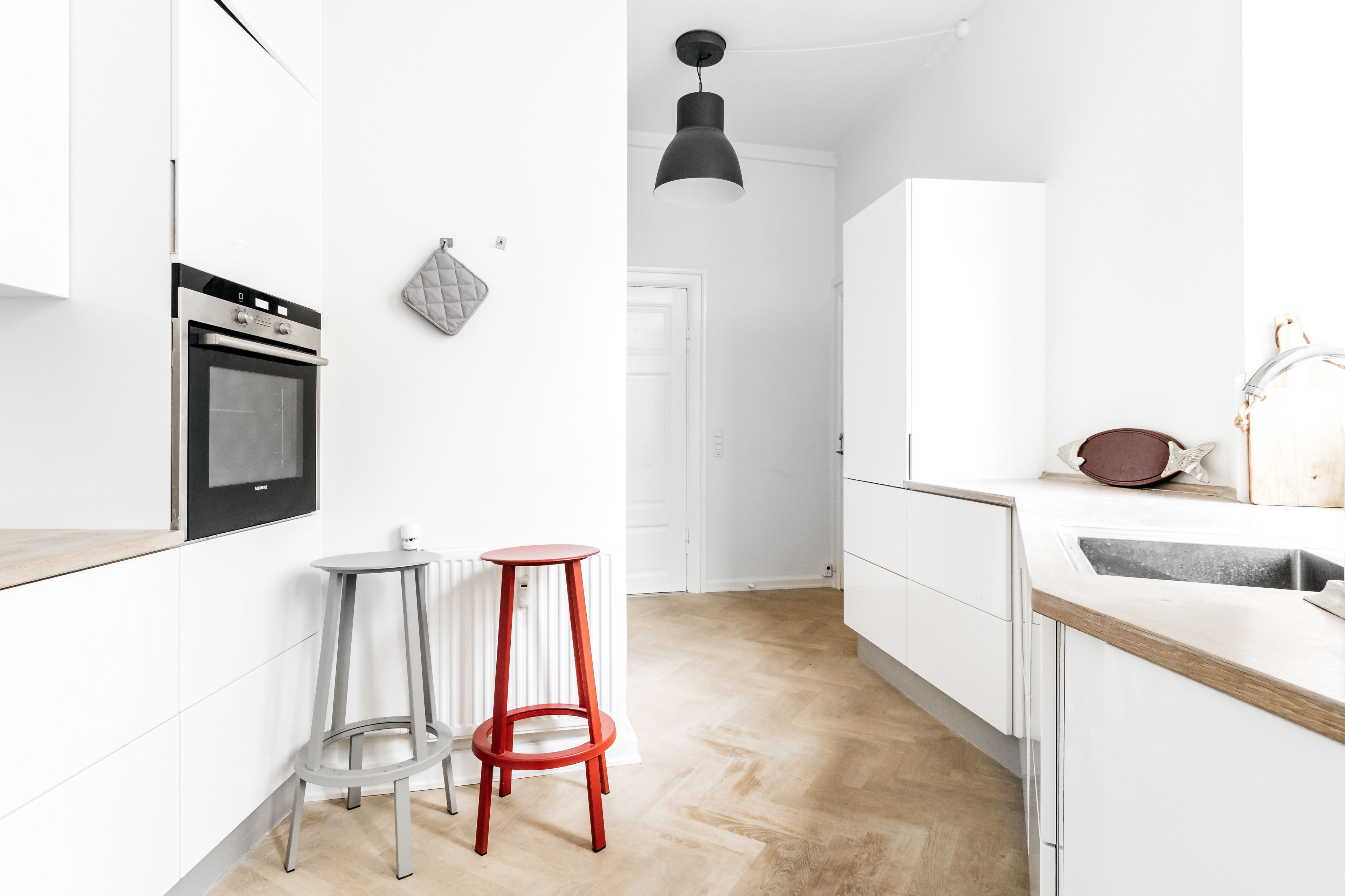 A modern kitchen with white cabinetry, a built-in oven, two stools, and a pendant light.