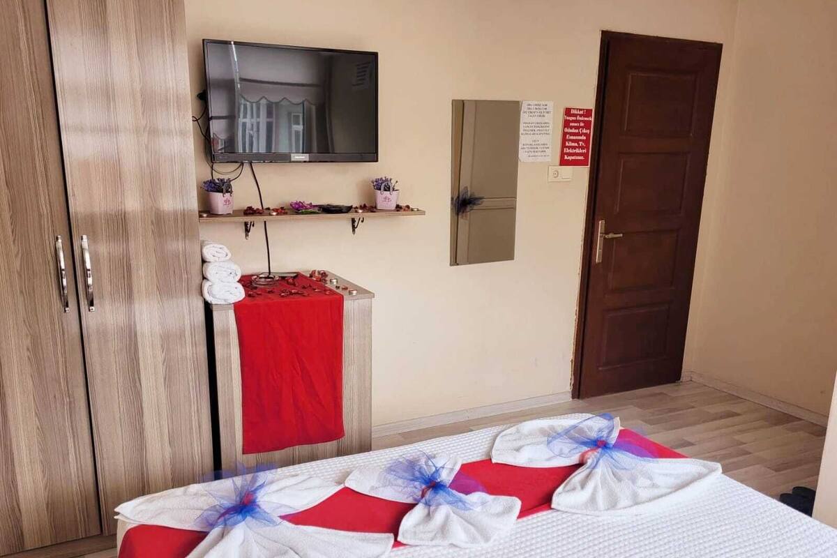 A hotel room with a bed, a TV mounted on the wall, a wooden wardrobe, a red table with a towel, and a door with a sign.