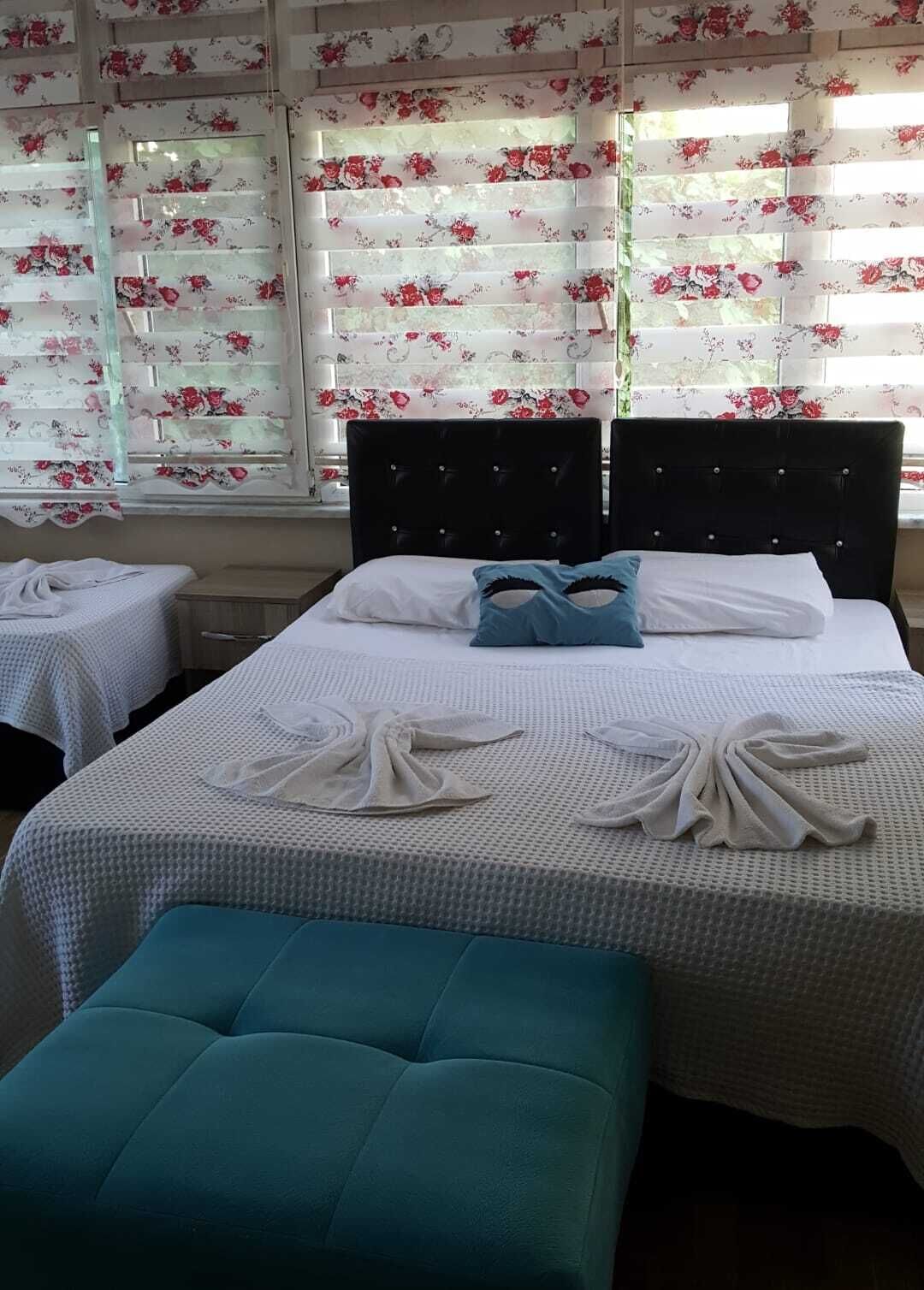 Habitación de hotel con dos camas, cada una con una colcha blanca y almohadas azules. Hay dos mesitas con manteles blancos, y la habitación tiene cortinas con motivos florales.