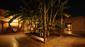 A well-lit courtyard with a stone pathway, surrounded by lush tropical plants and palm trees. - La Parenthese de Marrakech (Tameslouht)