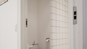 Et badeværelse med hvide flisemur, toilet, skraldespand og en dør til et andet rum.