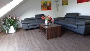 Ein modernes Wohnzimmer mit einem Ecksofa, einem Couchtisch und einer Topfpflanze. An den Wänden hängen zwei Bilder und darüber ein Oberlicht.