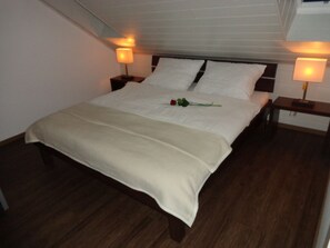 Una cama bien hecha con edredón blanco, dos almohadas y una sola rosa sobre la cama. Hay dos mesitas de noche con lámparas a cada lado de la cama.