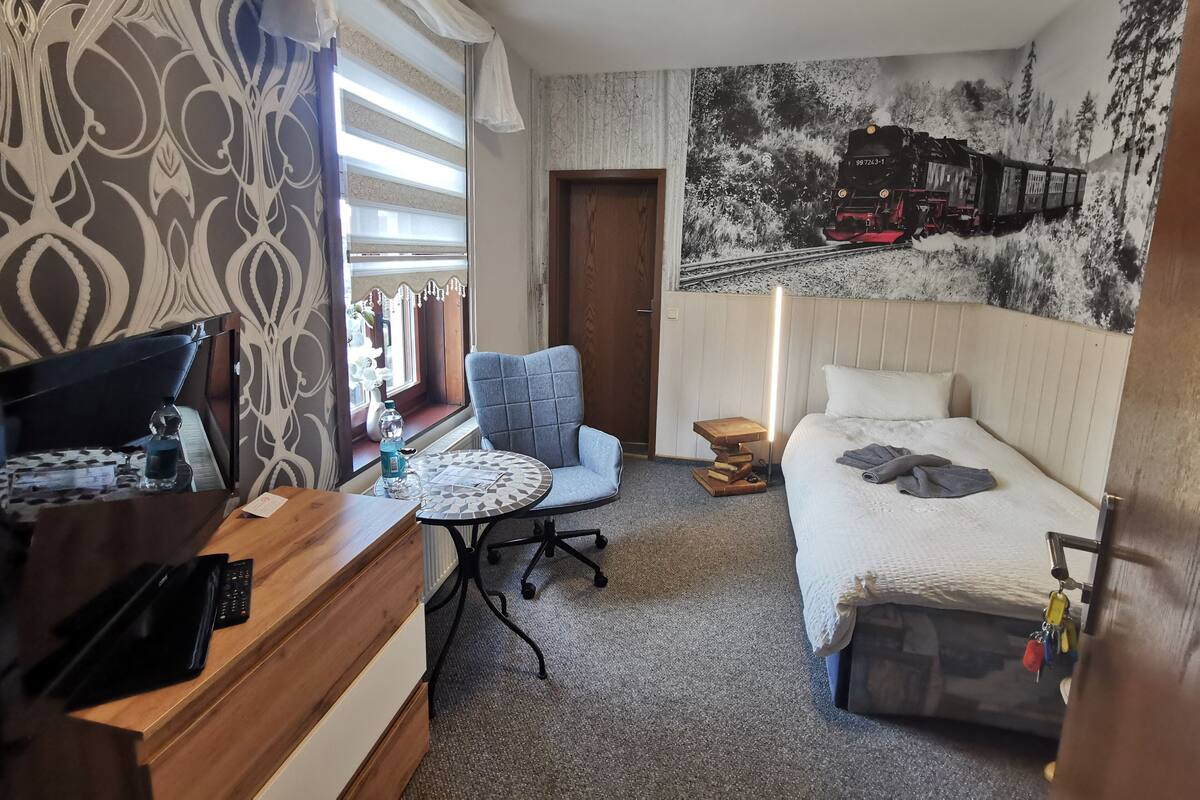 A hotel room with a bed, a desk with a computer, a chair, a small table, and a window with curtains.