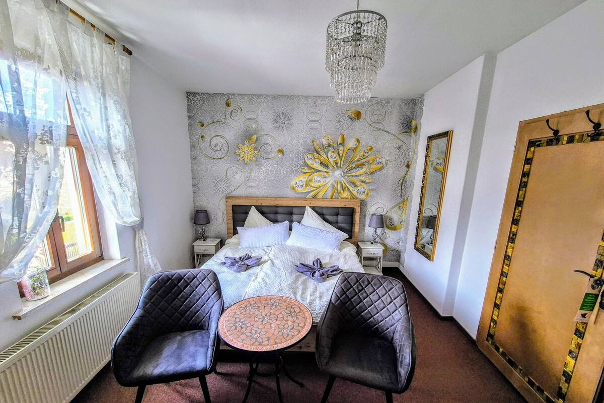 A bedroom with a bed, two chairs, a table, a chandelier, a window with curtains, and a decorative wall piece.