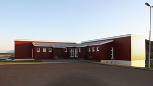 A red building with a flat roof, a central entrance, and a parking lot in front. - Framtid Hostel (Djupivogur)