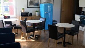 A compact kitchen with a blue refrigerator, a microwave, and a toaster. There are two dining tables with chairs, and a window with curtains. - Framtid Hostel (Djupivogur)