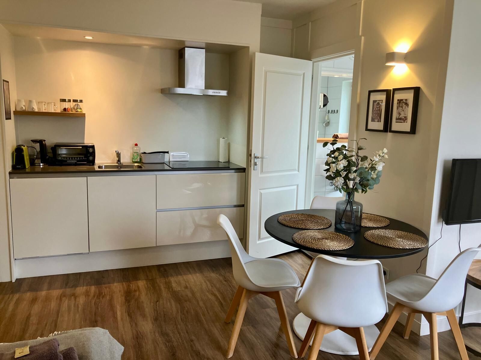 A modern kitchen with white cabinets, a toaster, and a coffee maker. There is a dining area with a round table, white chairs, and a vase with flowers.