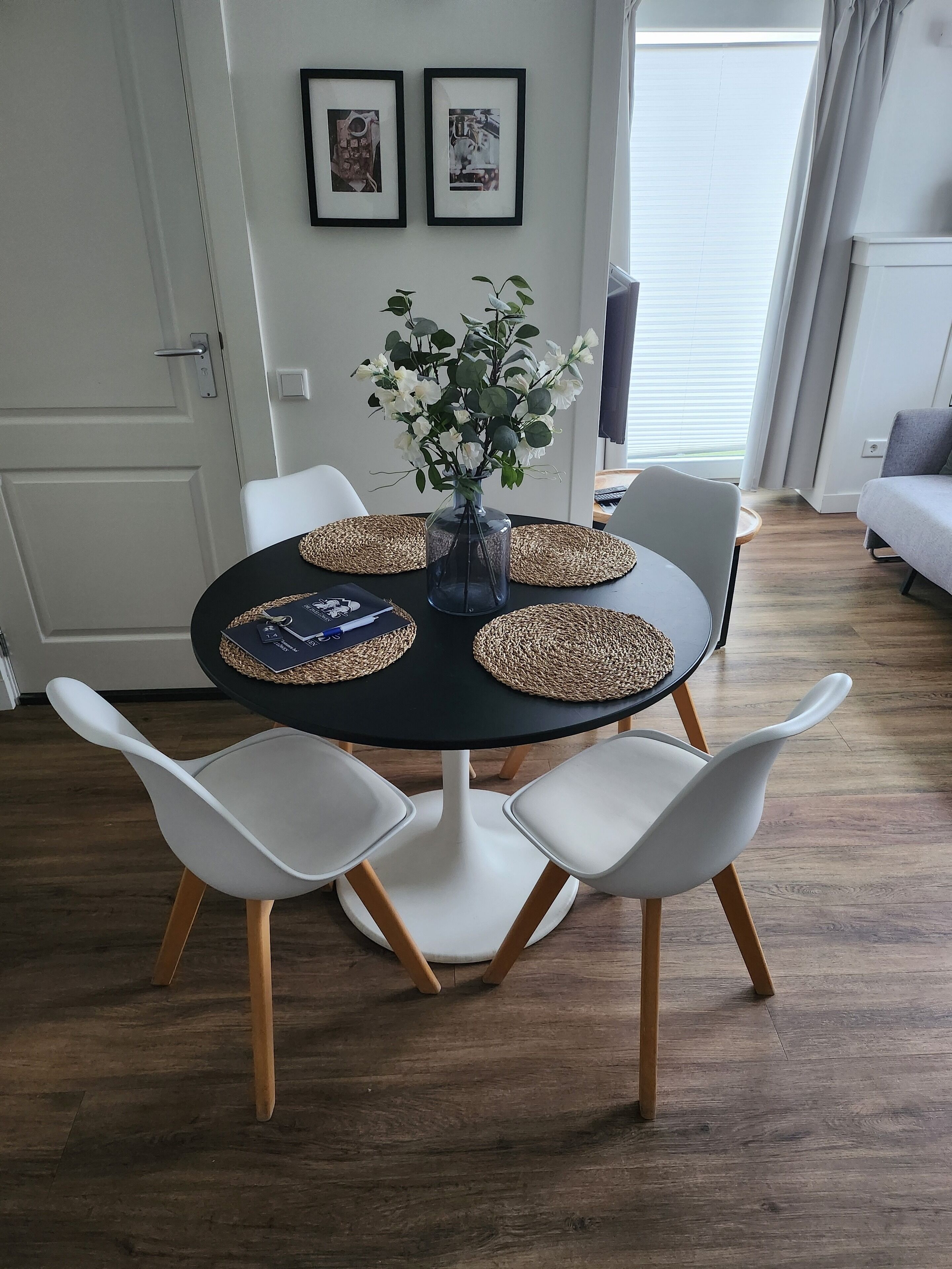 A dining area with a round table, white chairs, a vase with flowers, and two framed pictures on the wall.