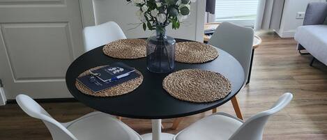 A dining area with a round table, white chairs, a vase with flowers, and two framed pictures on the wall.