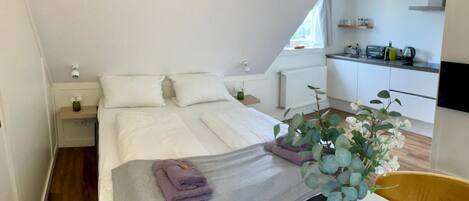A bedroom with a bed, bedside tables, a nightstand, a window, a plant, and a view of a kitchen.