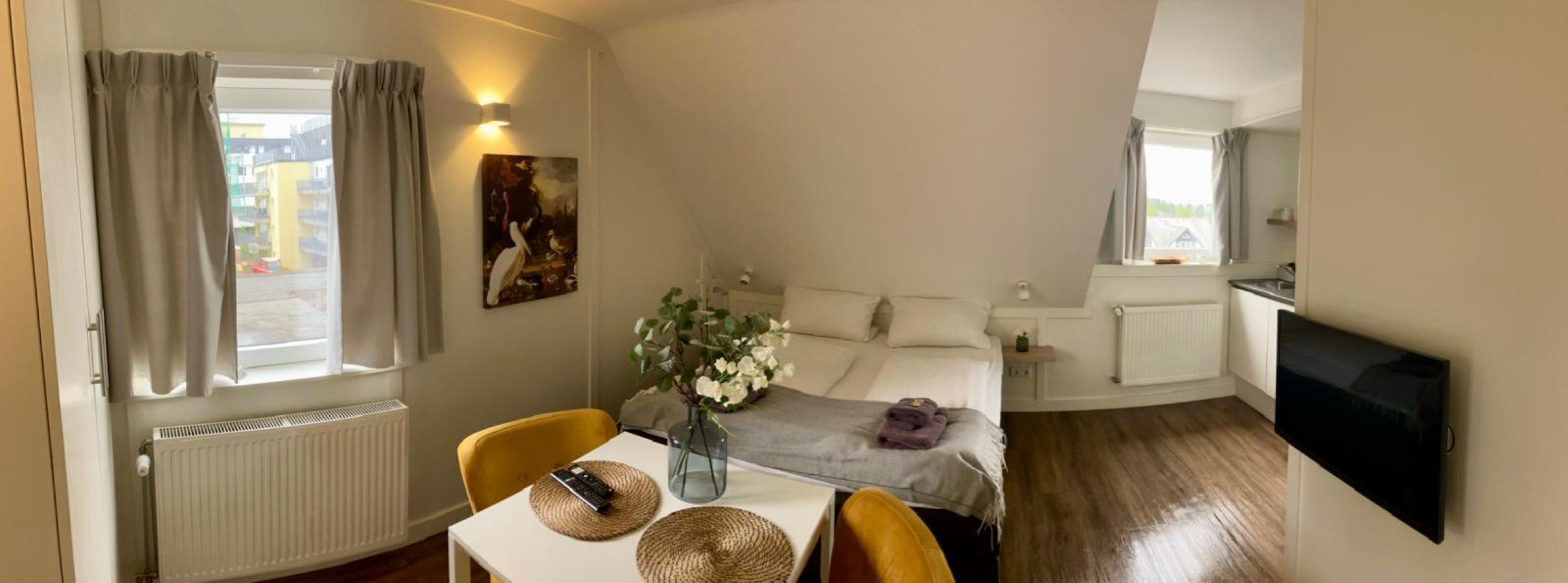 A hotel room with a bed, a desk with a vase of flowers, a window with curtains, a painting on the wall, and a TV mounted on the wall.