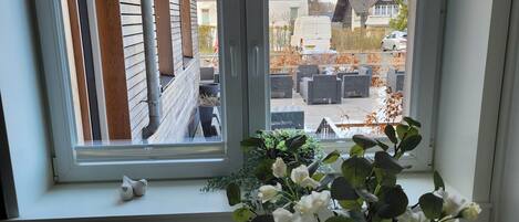 Ein Essbereich mit Tisch für zwei Personen, einem Fenster mit Blick auf eine Wohnstraße und einer Vase mit weißen Blumen.
