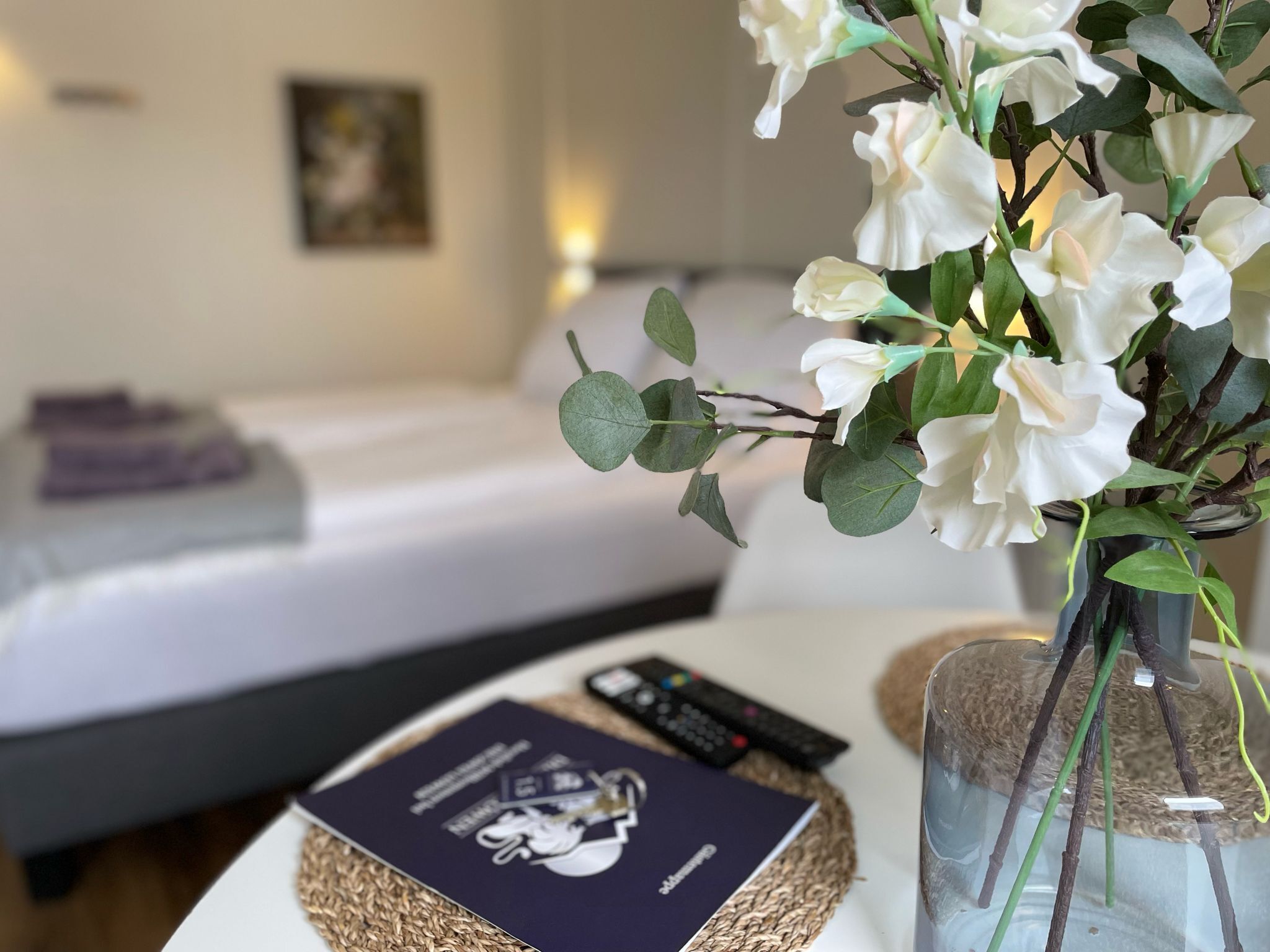 A neatly made bed with a purple towel, a remote control, a book, and a vase with white flowers on a bedside table.