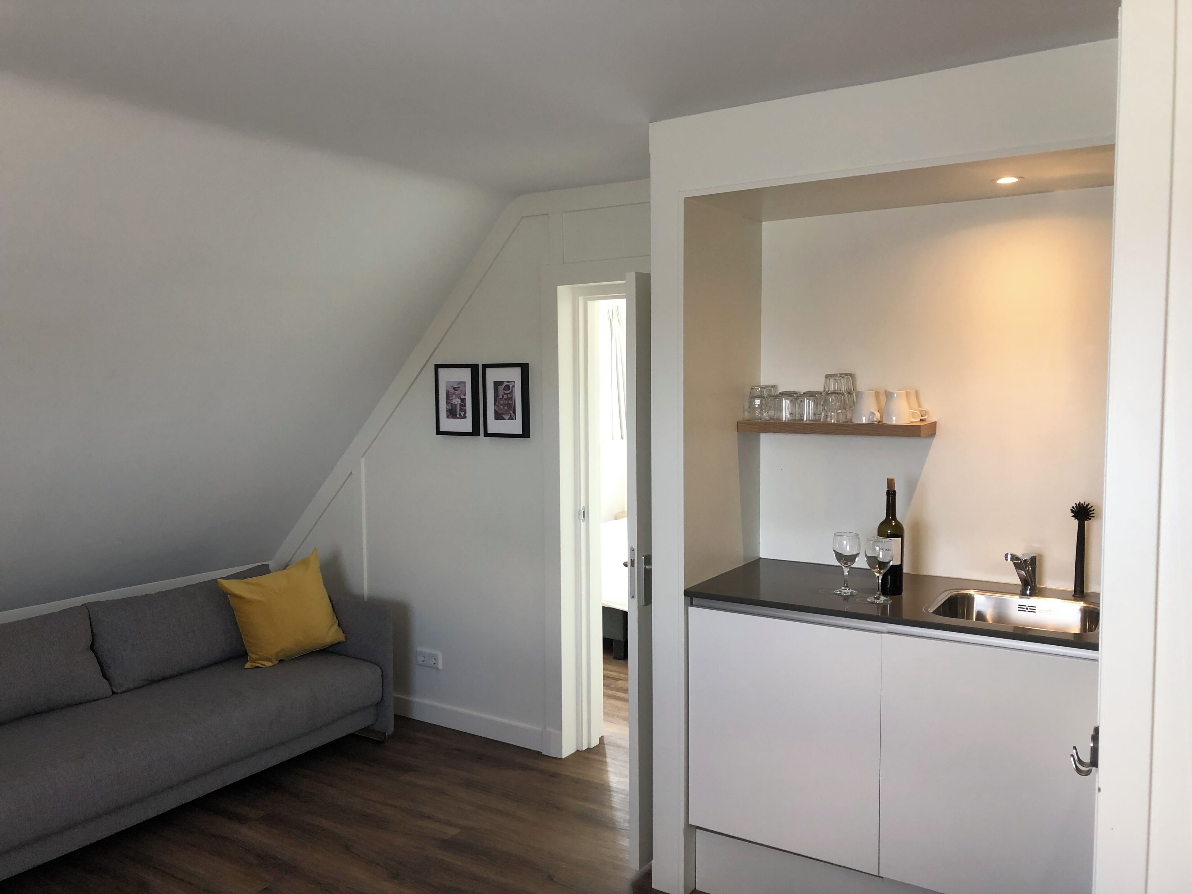 Un espacio habitable compacto con kitchenette, un sofá gris con un cojín amarillo y un área de comedor con una estantería que contiene vasos y una botella.
