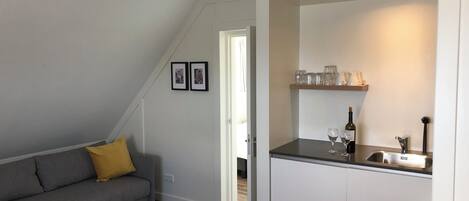 Un espacio habitable compacto con kitchenette, un sofá gris con un cojín amarillo y un área de comedor con una estantería que contiene vasos y una botella.