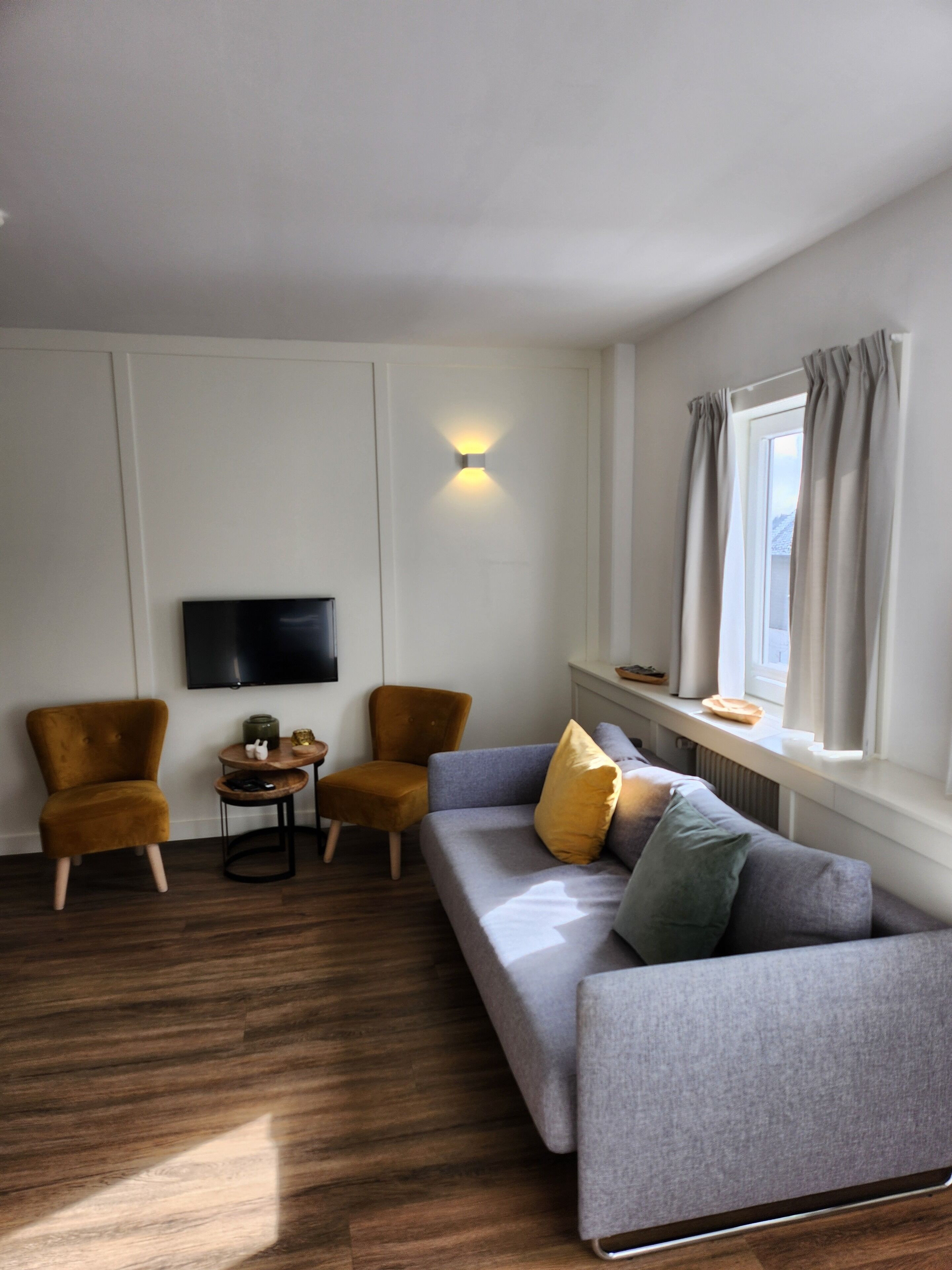A modern living room with a grey sofa, two armchairs, a small table with decorative items, a flat-screen TV mounted on the wall, and a window with curtains.