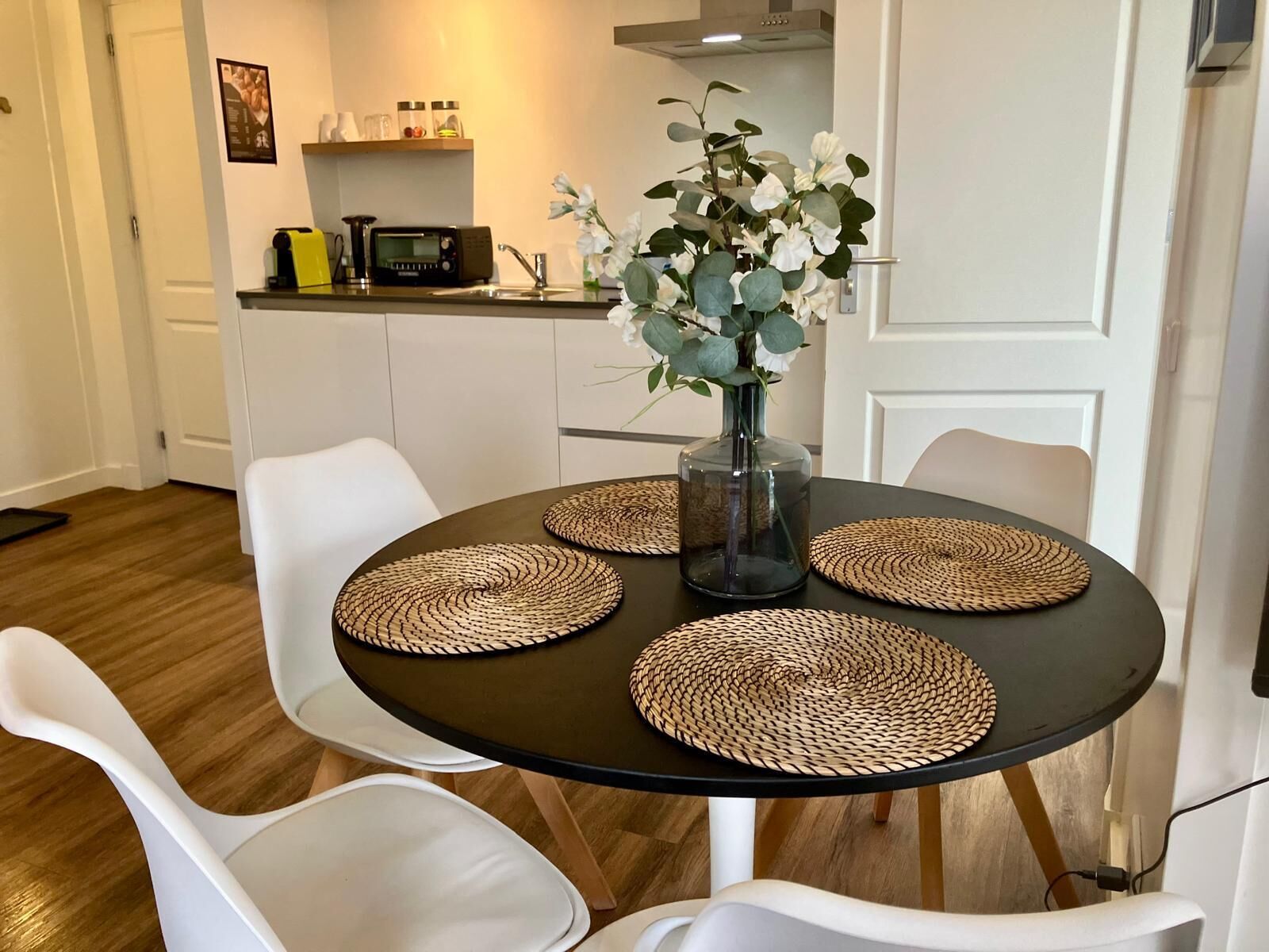 A dining area with a round table, four woven placemats, a vase with white flowers, and white chairs.