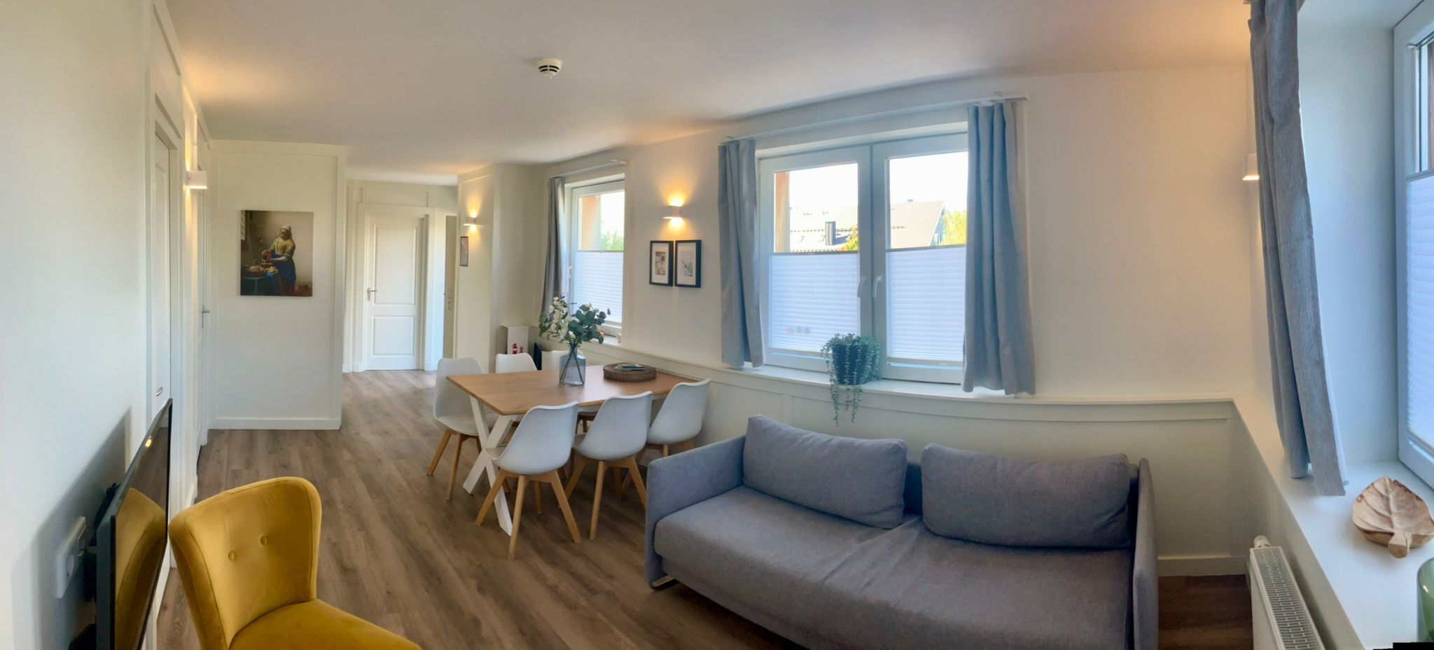 A modern living room with a sofa, dining table, and chairs. There are windows with curtains and a view of the outside.