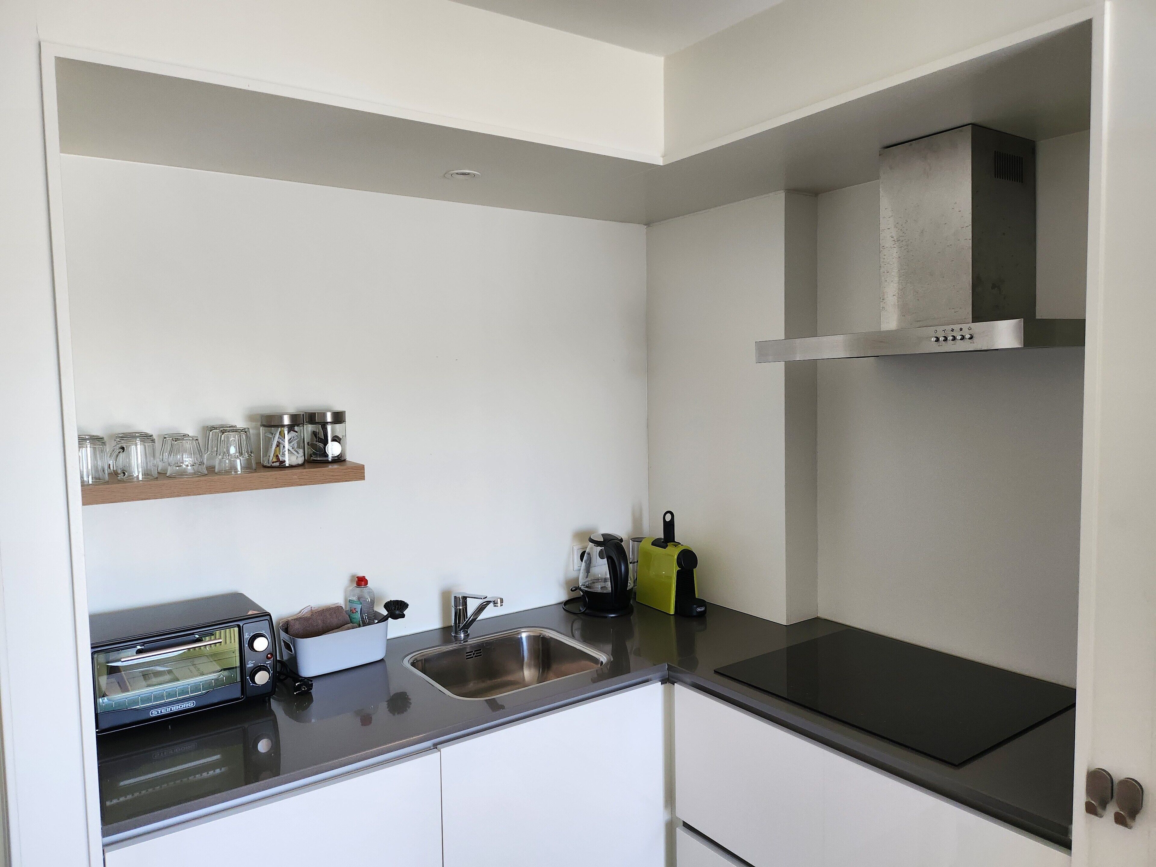 Una cocina moderna con horno empotrado, pileta de acero inoxidable y campana extractora. Hay frascos de vidrio en una estantería y un horno tostador en la encimera.