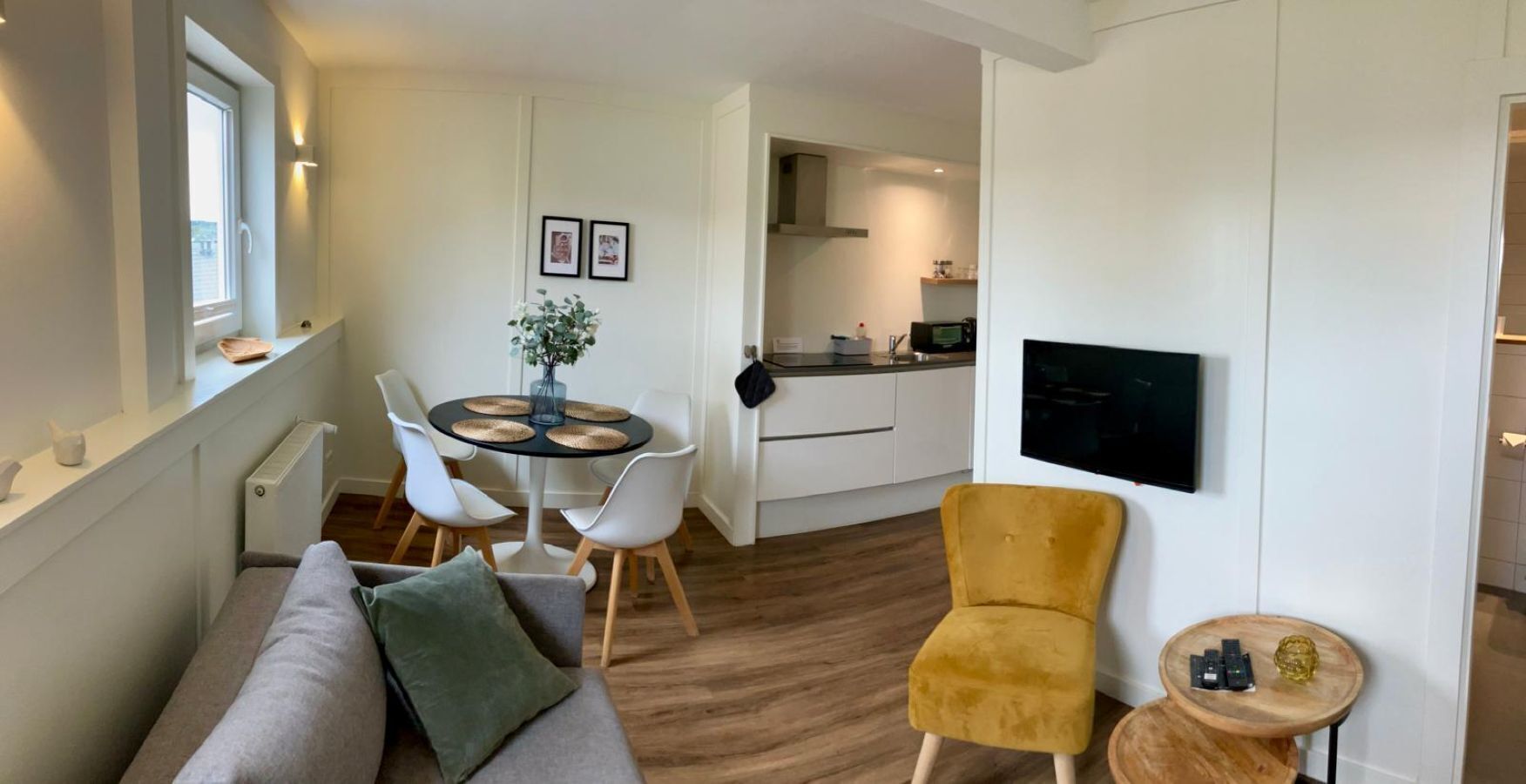 Una sala de estar moderna con un sofá, un sillón amarillo, una mesa de comedor con sillas, una pequeña cocina y un televisor montado en la pared.