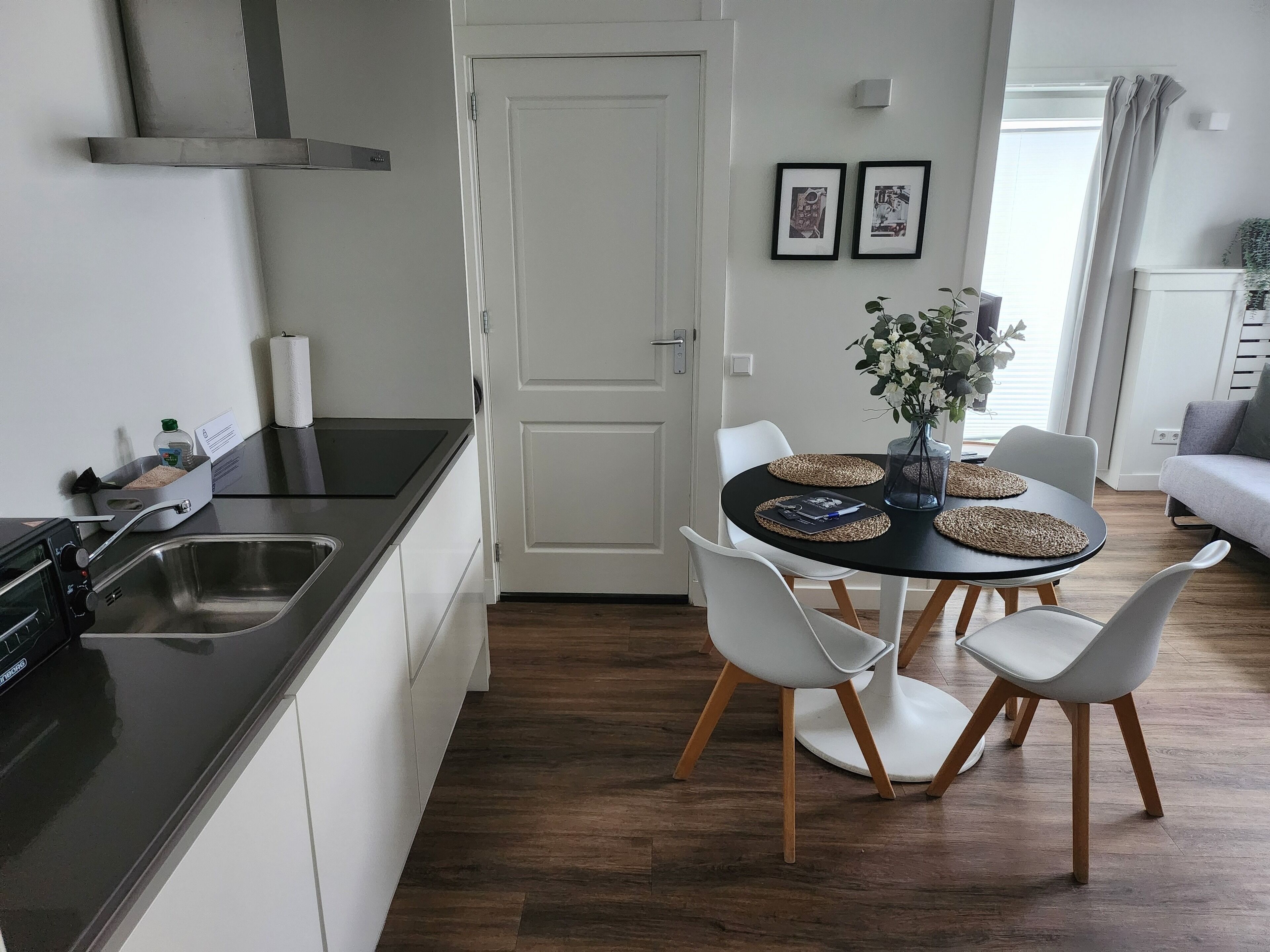 Una cocina moderna con encimera negra, armarios blancos y pileta de acero inoxidable. Hay un comedor con mesa redonda, sillas blancas y un florero con flores.